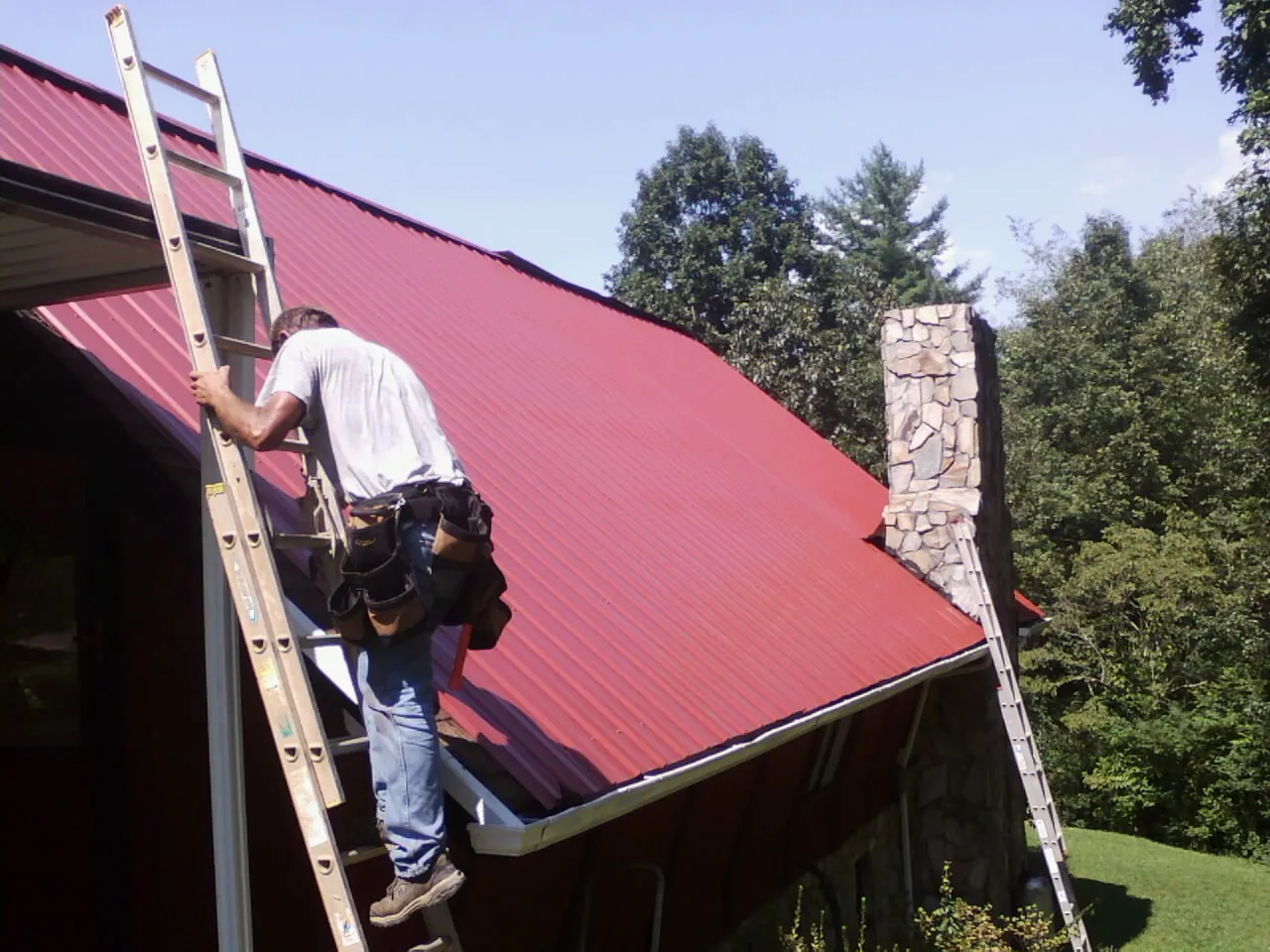 Roofer on a ladder beside a red metal roof and stone chimney — Soffit & Fascia Repair by Roofing Brothers in Butte City